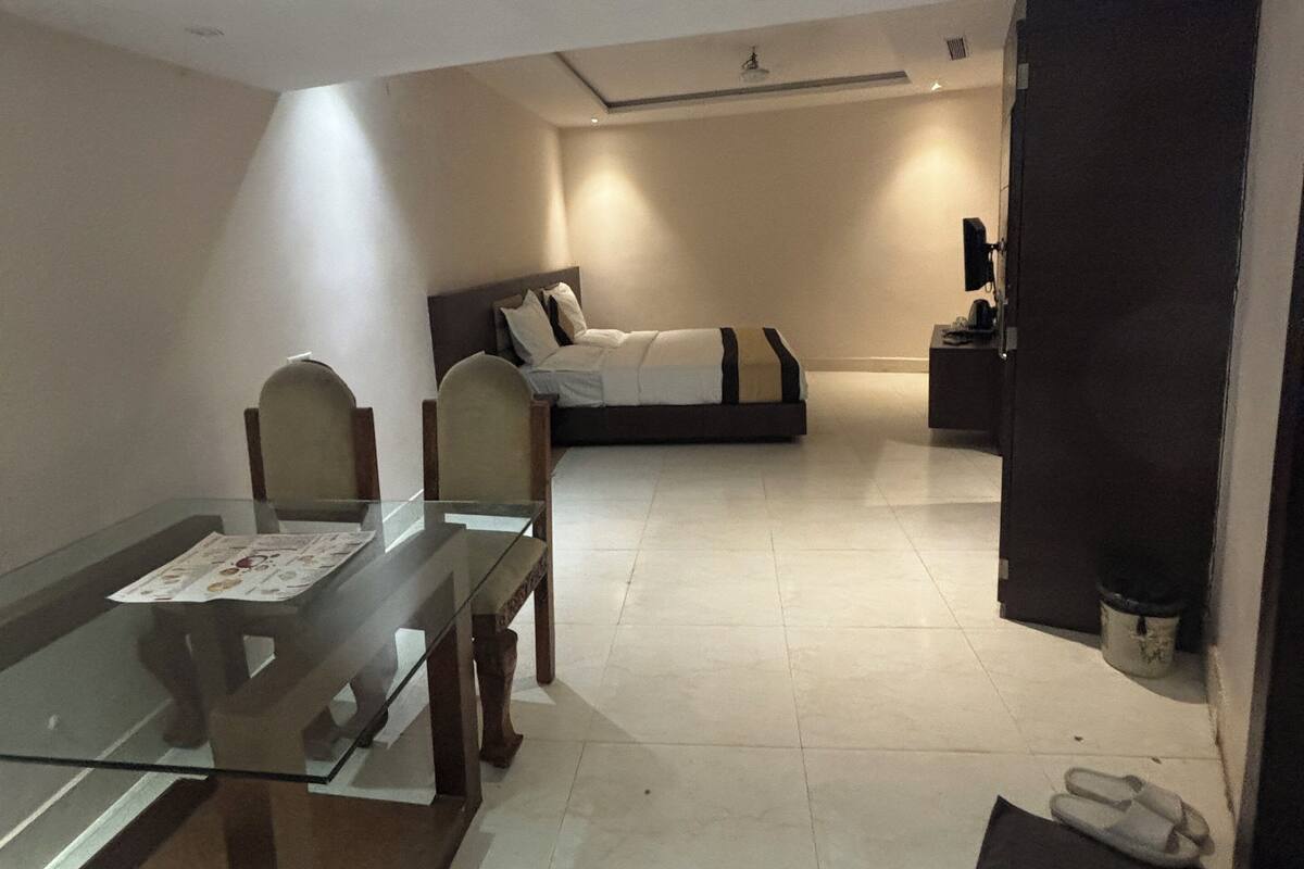 A hotel room with a glass dining table, two chairs, a bed, a bedside table, and a small TV.