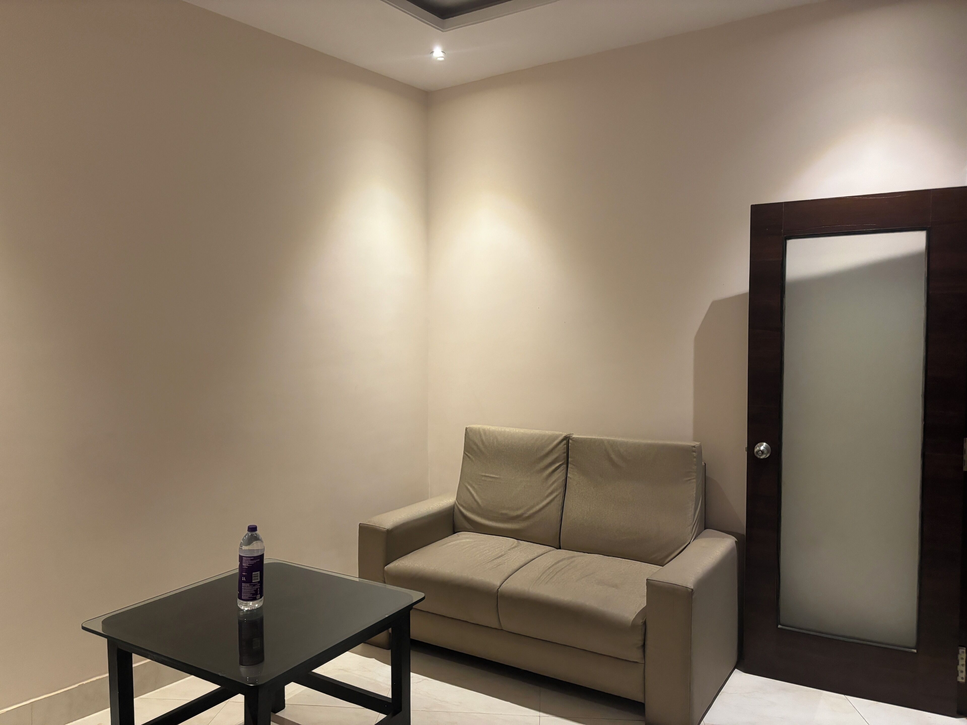 Una habitación pequeña y moderna con un sofá color beige, una mesa con superficie de vidrio y una puerta con paneles de vidrio esmerilado.