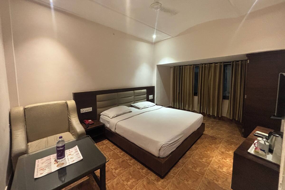 A hotel room with a bed, a small table with a bottle, a chair, a nightstand, and a TV.