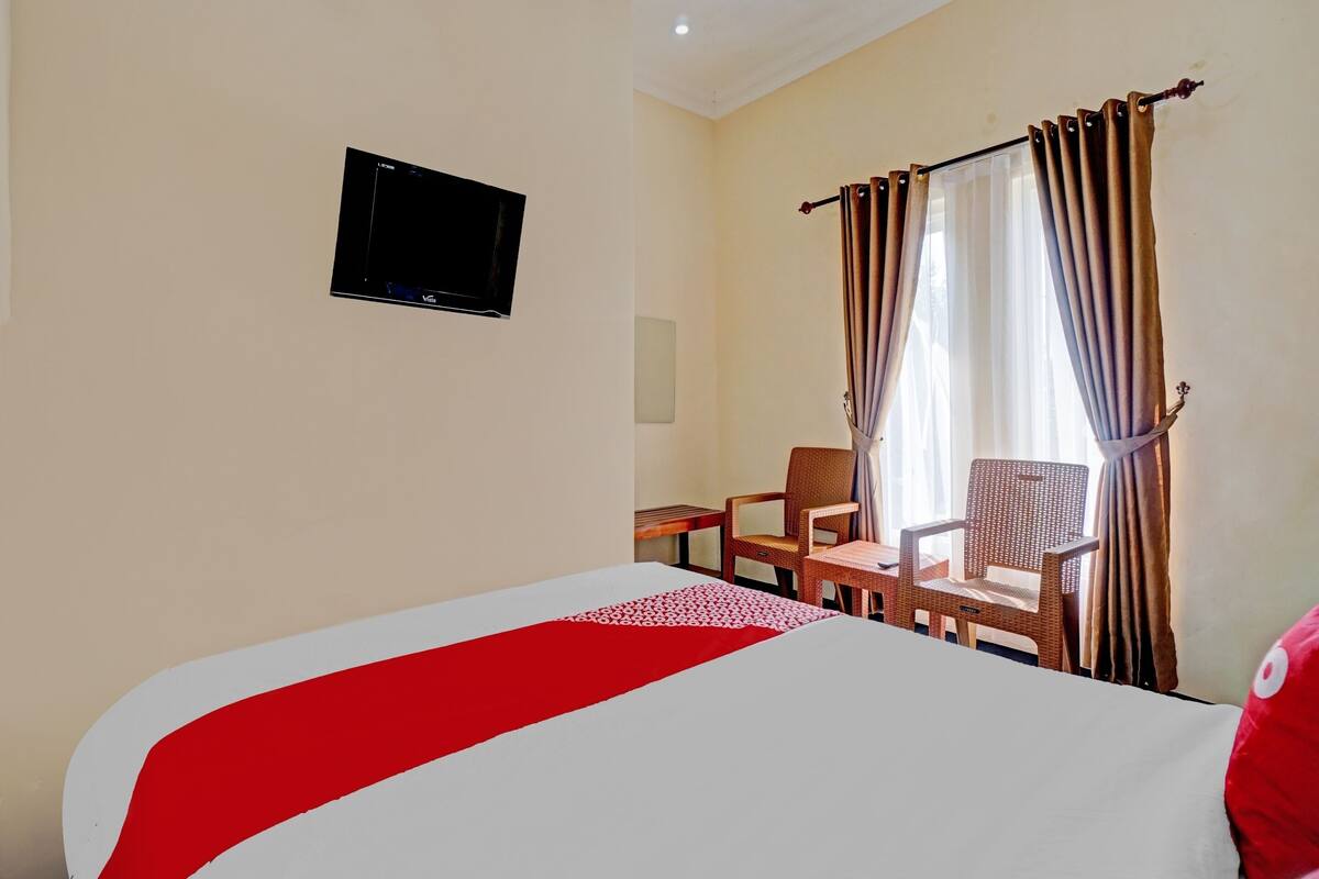 Quarto de hotel com uma cama, televisão, escrivaninha, duas cadeiras e uma janela com cortinas.