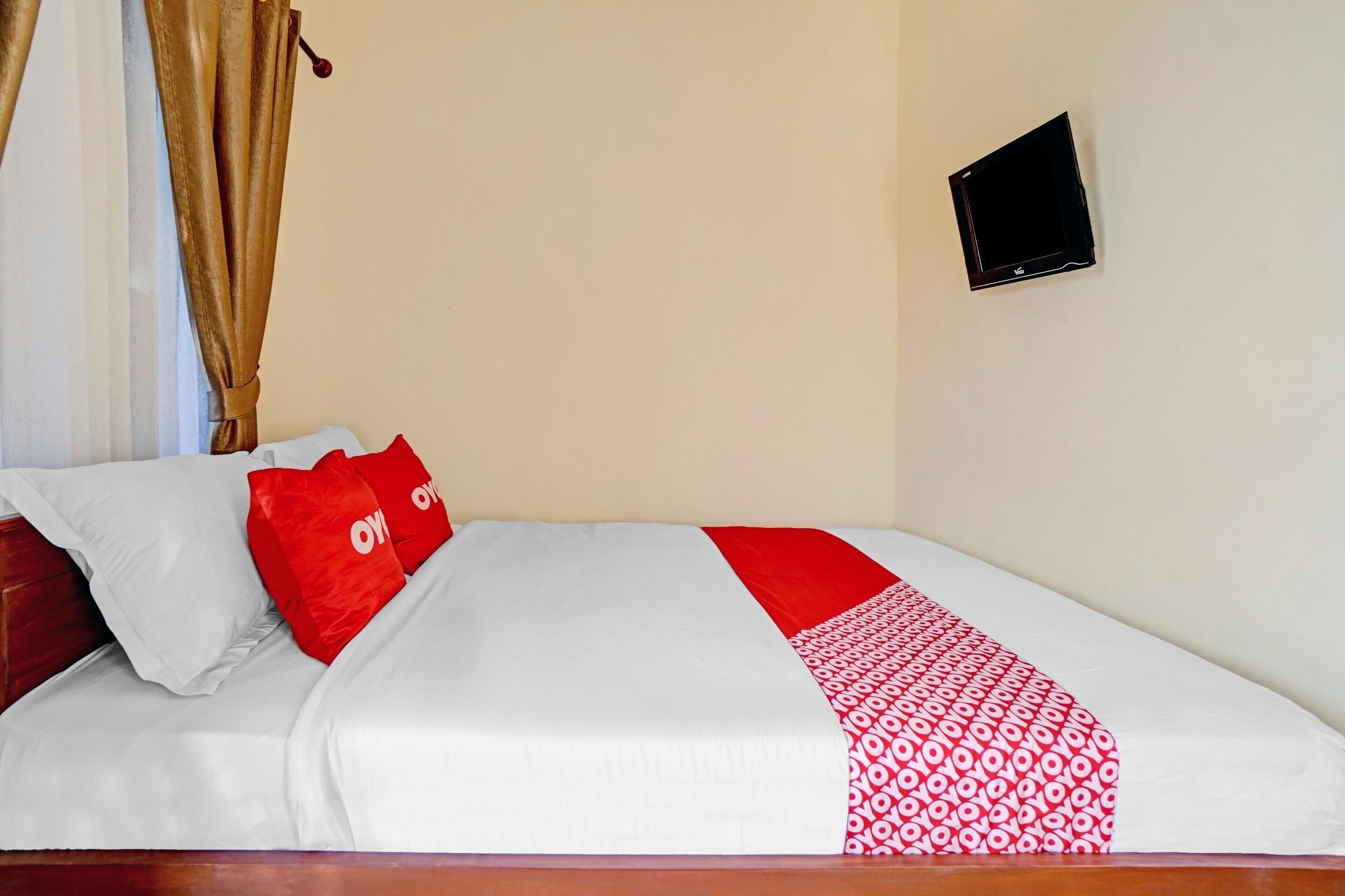 Uma cama com roupa de cama branca e vermelha, um travesseiro vermelho e uma TV fixada na parede.