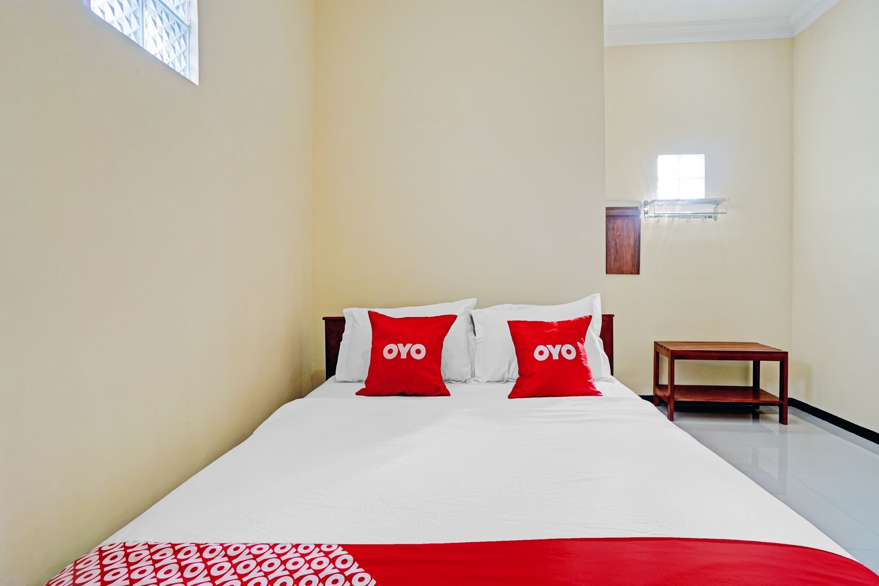 Une chambre d’hôtel avec un lit, deux oreillers rouges ornés du logo « OYO », une table de chevet en bois et une fenêtre avec des stores.