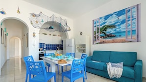 A bright living room with a teal sofa, a dining area with blue chairs, and a kitchen in the background. - Casa Celeste 100m From Beach (Porto Cesareo)