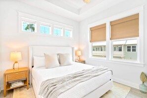 Un dormitorio con una cama grande, dos mesitas de noche con lámparas, una ventana con persianas y vistas a un edificio.