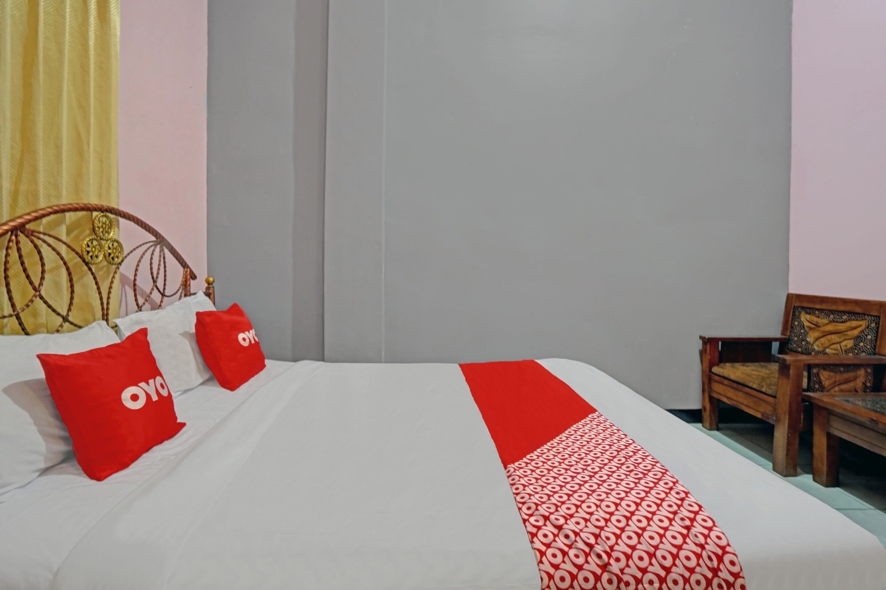 A neatly made bed with a white and red bedspread, two red pillows with the word "OYO" on them, and a wooden chair with a cushion in the corner.