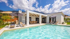 A modern poolside area with a clear pool, outdoor seating, and a mountain backdrop. - Villa Petra in Afrato (Kefalonia)