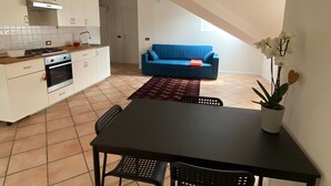 Una cocina compacta con gabinetes blancos y una estufa a gas, un sofá azul en la sala de estar, y una mesa de comedor con sillas.