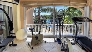 A home gym with exercise equipment, a large window with a view of palm trees and a pool. - Villa 'Luxury Xenofilia Lll' with Mountain View, Wi-Fi and Air Conditioning (Moraira)