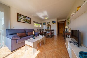 Uma sala de estar com sofá, mesa de centro e televisão. Há uma área de refeições com cadeiras e mesa.