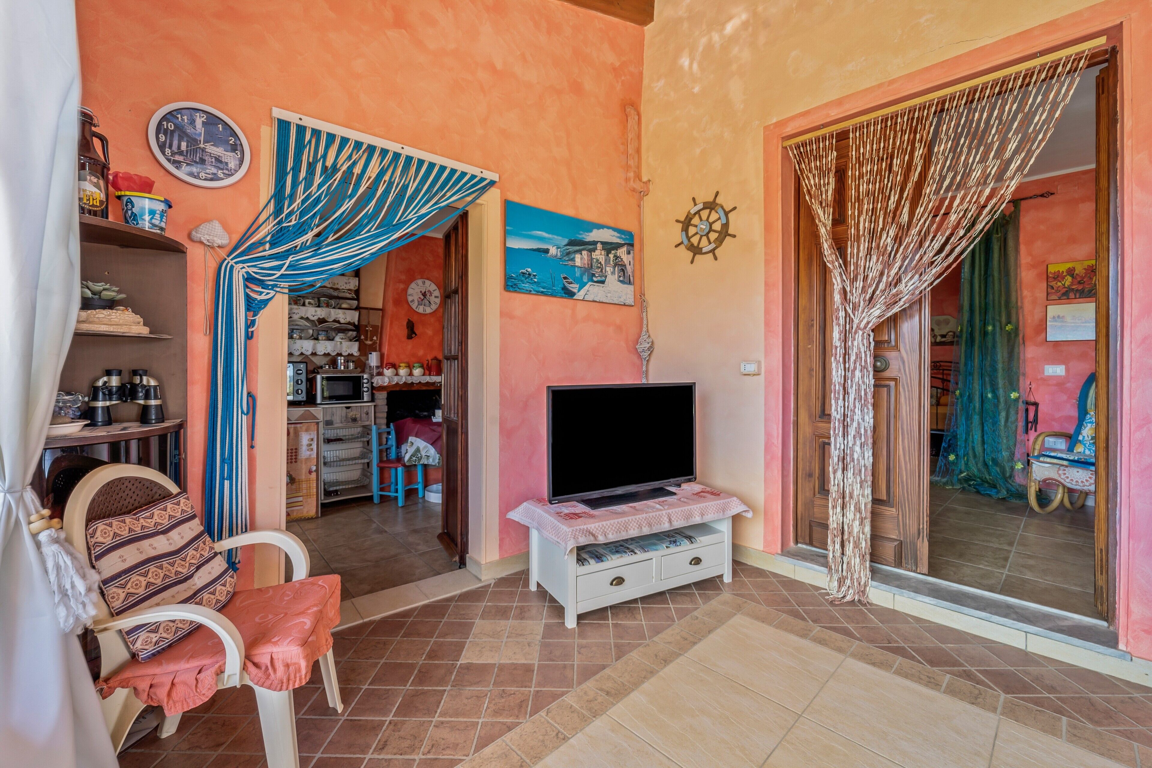 Una sala de estar con televisor, un sillón, un estante con utensilios de cocina y una puerta que da a otra habitación.