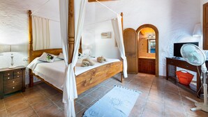 A four-poster bed with a canopy, a wooden desk with a fan, and a bathroom visible through an archway.