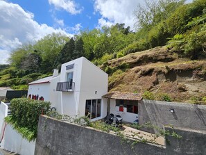 A white building with a balcony, a small attached structure with a tiled roof, and a view of a hillside. - Holiday Home 'Paradise House Furnas' with Mountain View, Private Terrace and Wi-Fi (Furnas)