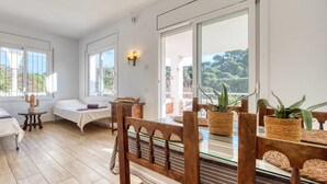 A room with two beds, a dining table with chairs, a cactus, and a view of trees through large windows.