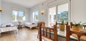 A room with two beds, a dining table with chairs, a cactus, and a view of trees through large windows.