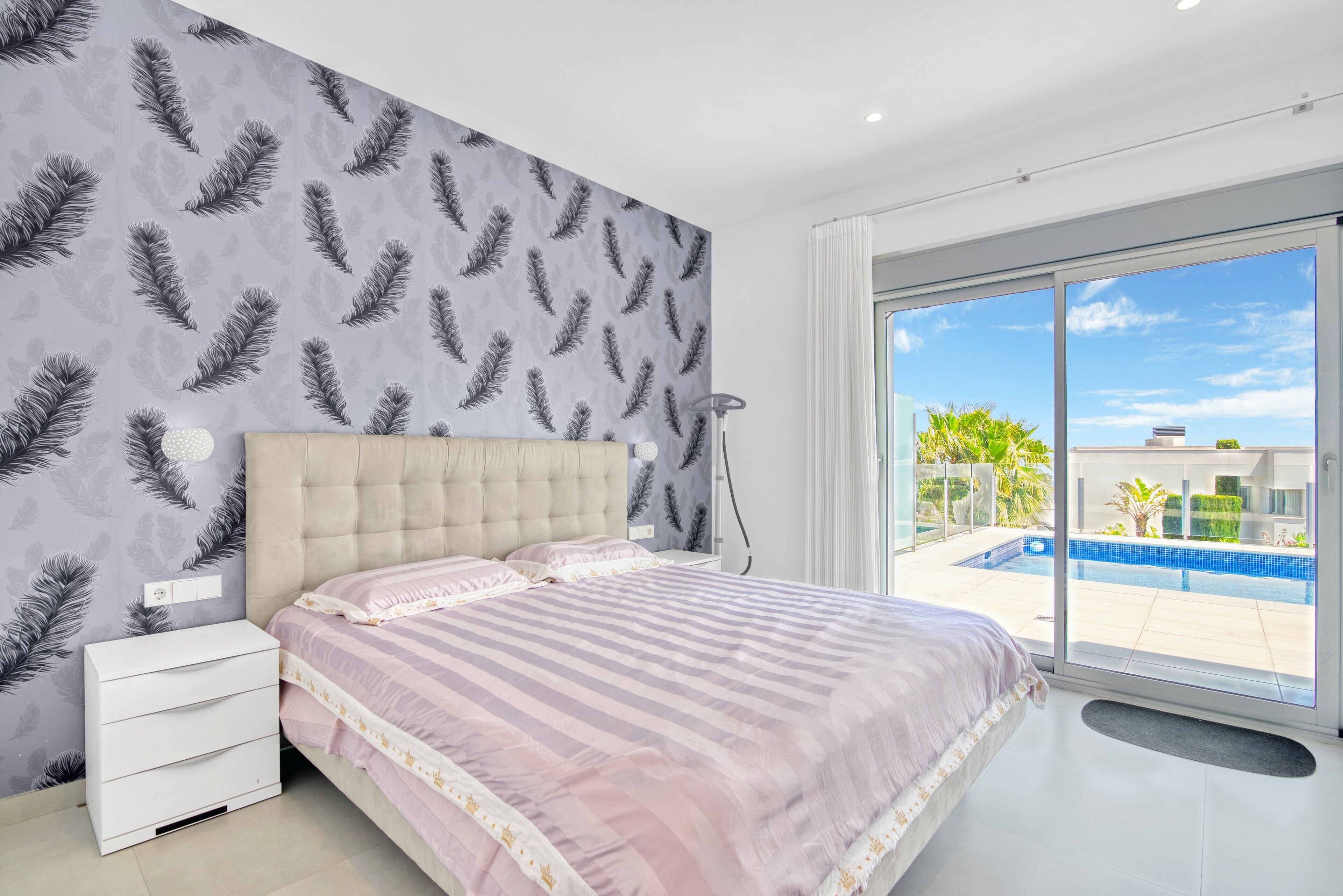 A bedroom with a large bed, a nightstand, a wall-mounted lamp, and a view of a swimming pool through a glass door.