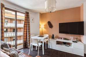 A modern living room with a sofa, a dining table, chairs, a TV, and a balcony with a view of a building.
