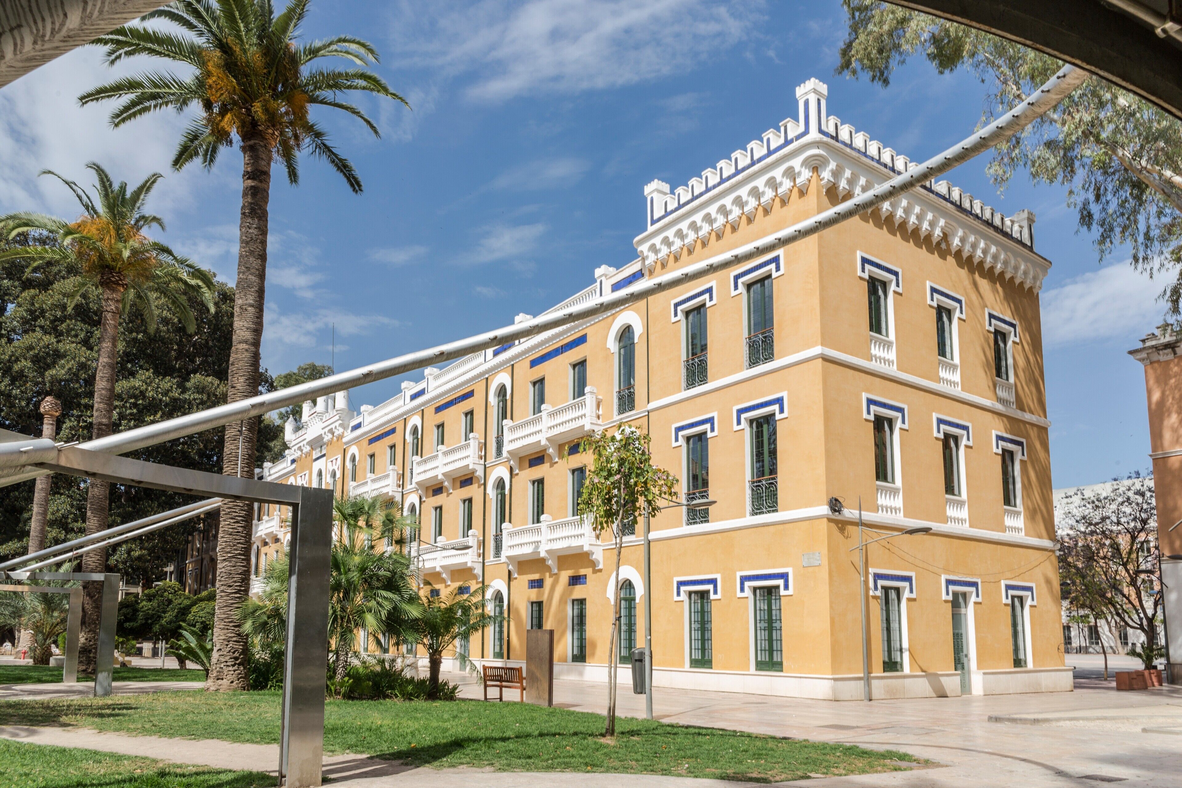 A yellow building with white trim and multiple windows, surrounded by palm trees and greenery.