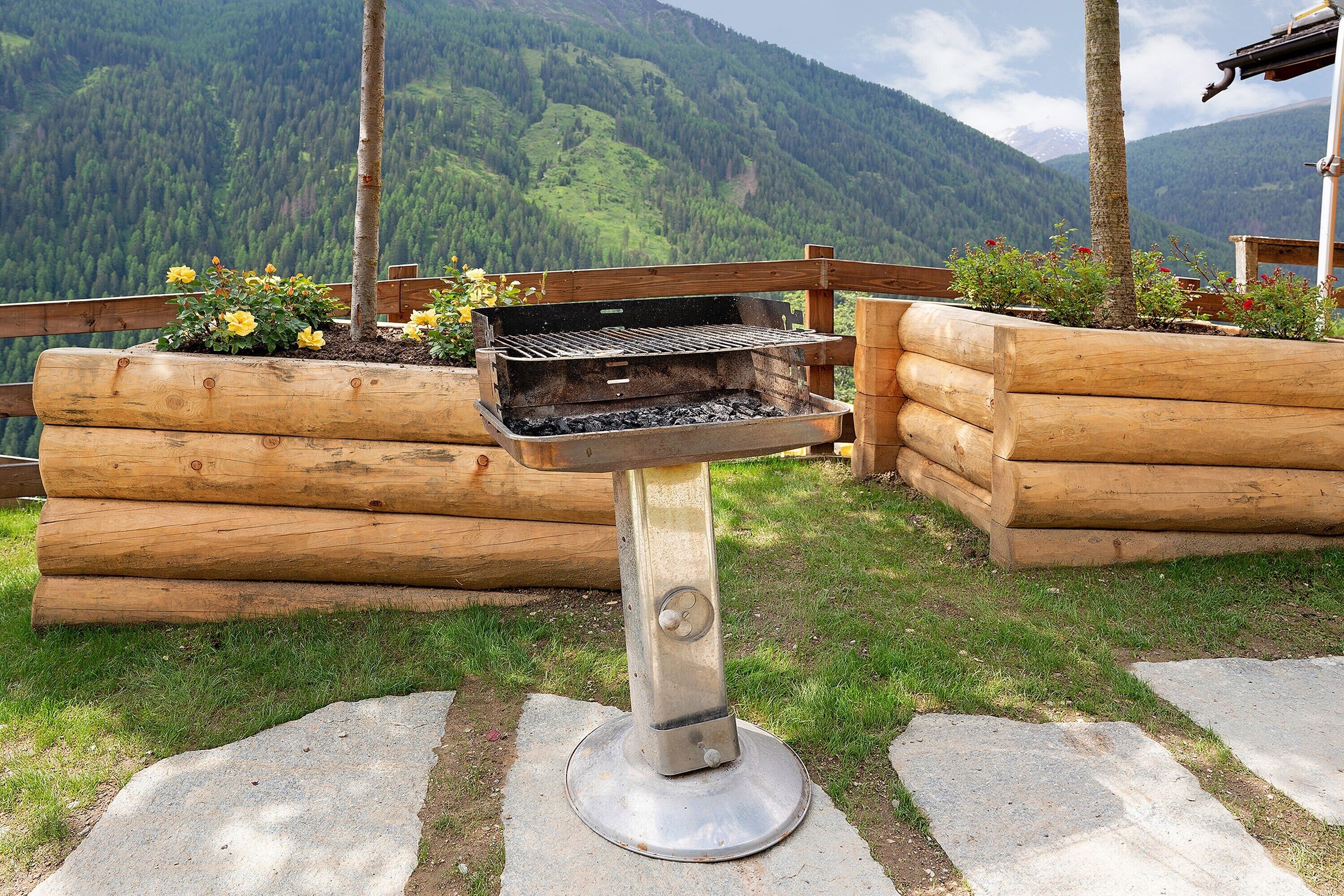 A grill with a metal stand, surrounded by wooden logs and a grassy area with flowers.