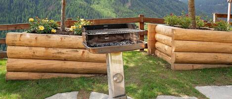 A grill with a metal stand, surrounded by wooden logs and a grassy area with flowers.