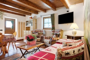 A cozy living room with wooden beams, a television, a sofa, a chair, a table with a vase of flowers, and a window with curtains.