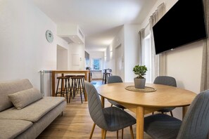 Uma sala de estar moderna com um sofá, uma mesa de jantar com cadeiras, uma televisão na parede e um vaso com planta na mesa.