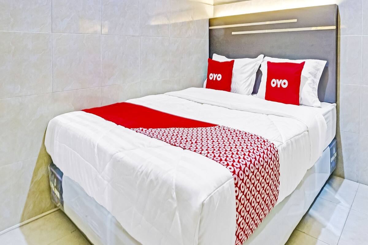 A hotel room with a bed, white and red bedding, and a headboard with a logo.