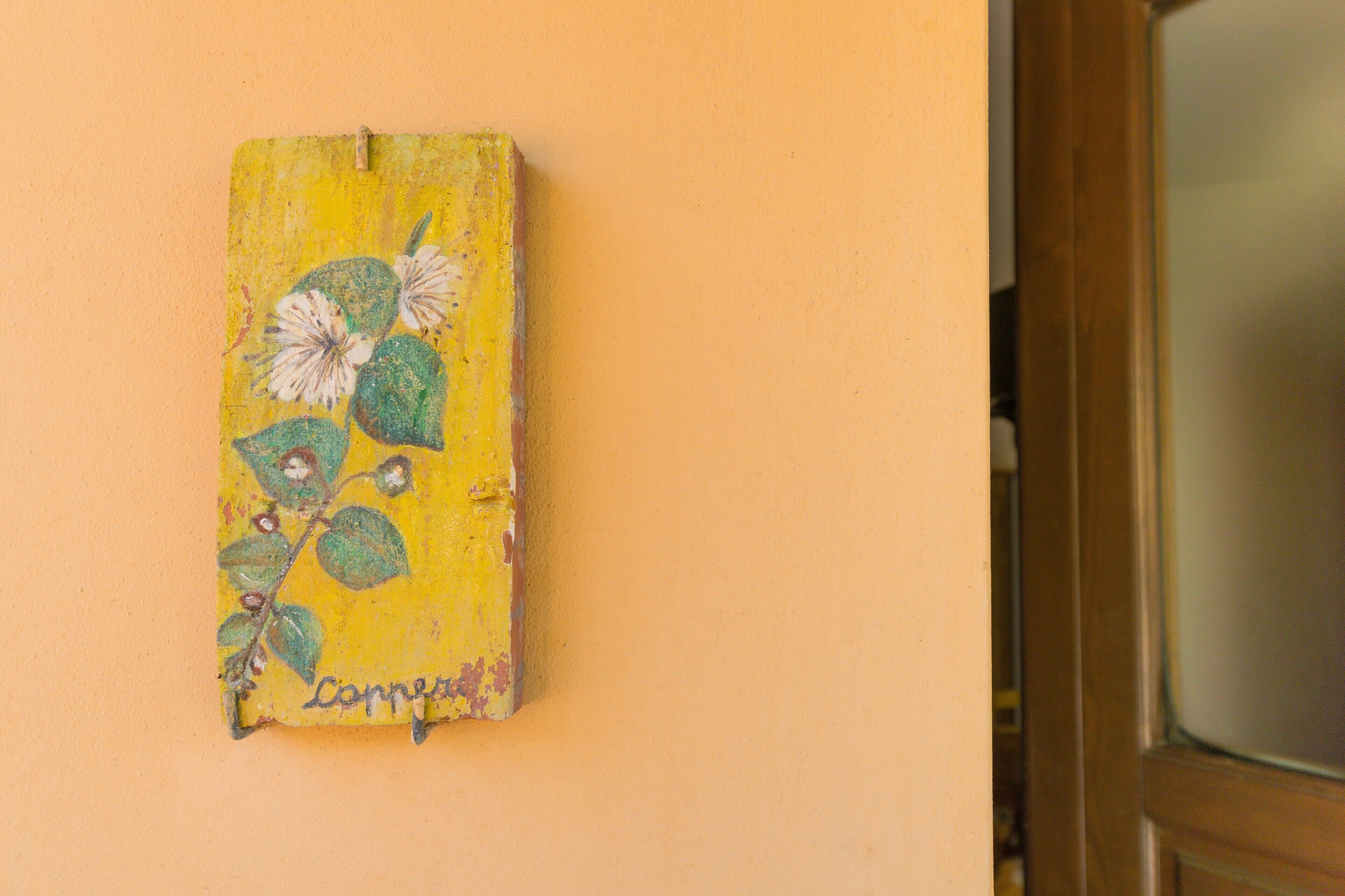 Un mur jaune orné d’un petit tableau usé représentant des fleurs et des feuilles.