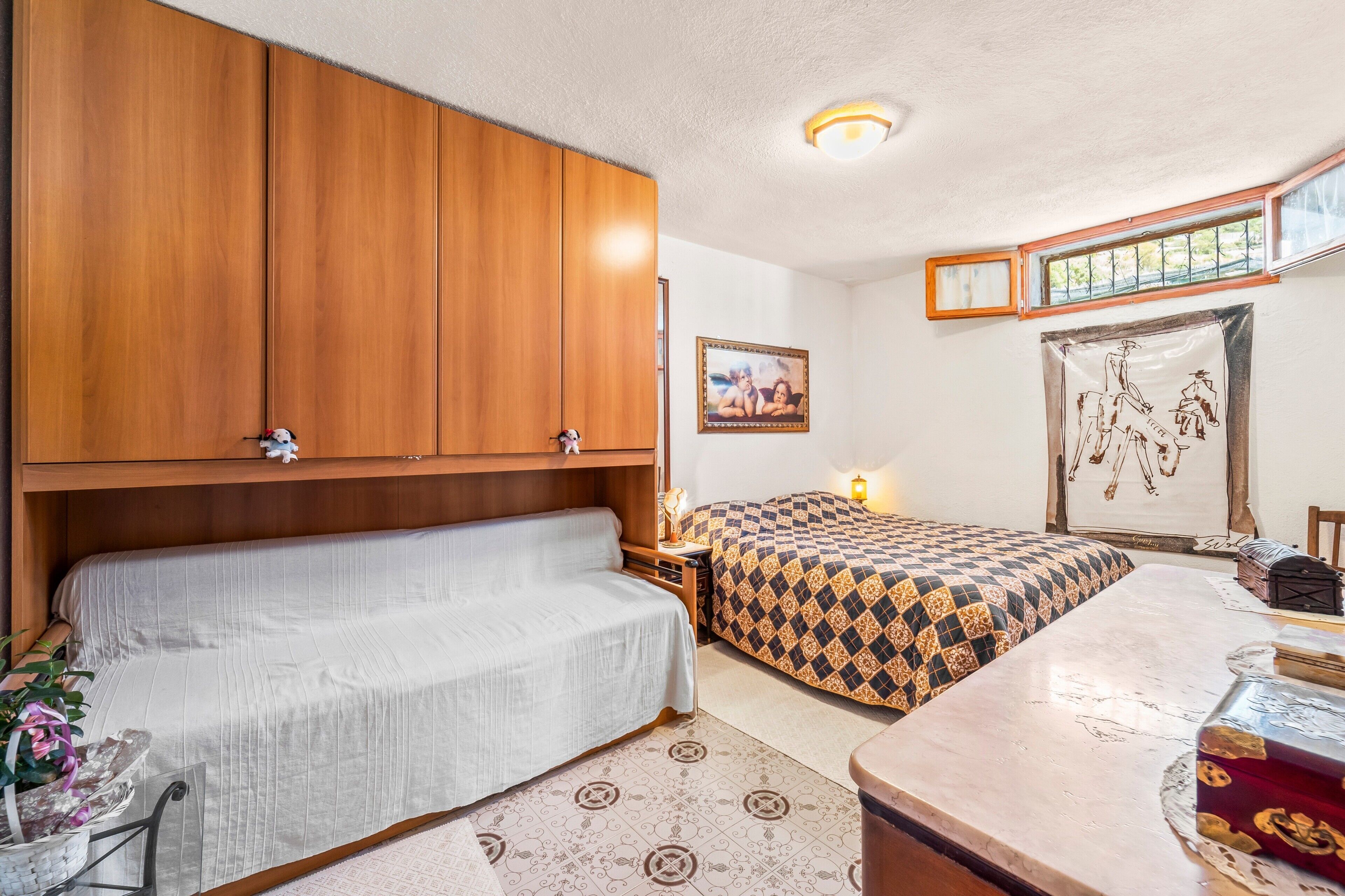 Une chambre avec une armoire en bois, un canapé, un lit avec une literie à motifs, une table de chevet, une lampe, une fenêtre donnant sur un paysage agréable et un tableau encadré au mur.
