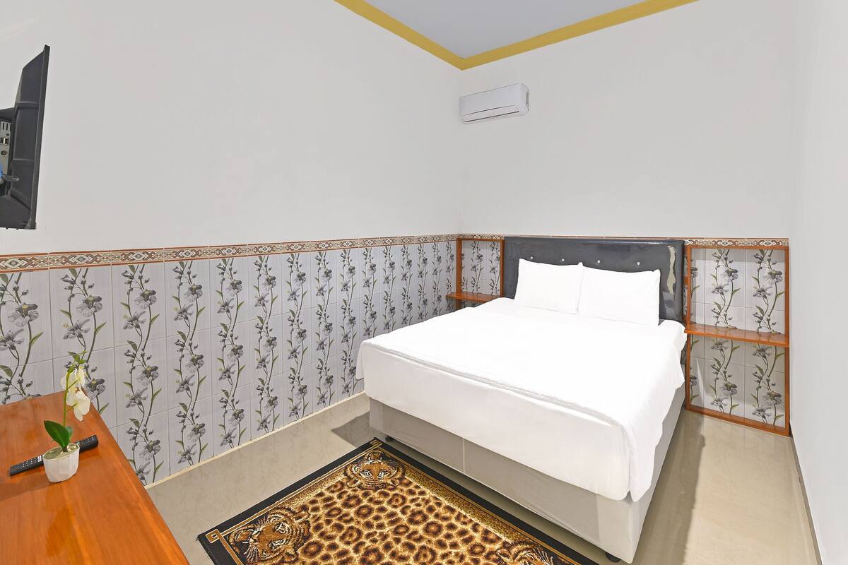 A bedroom with a bed, a desk, a chair, a small table, and a decorative plant.