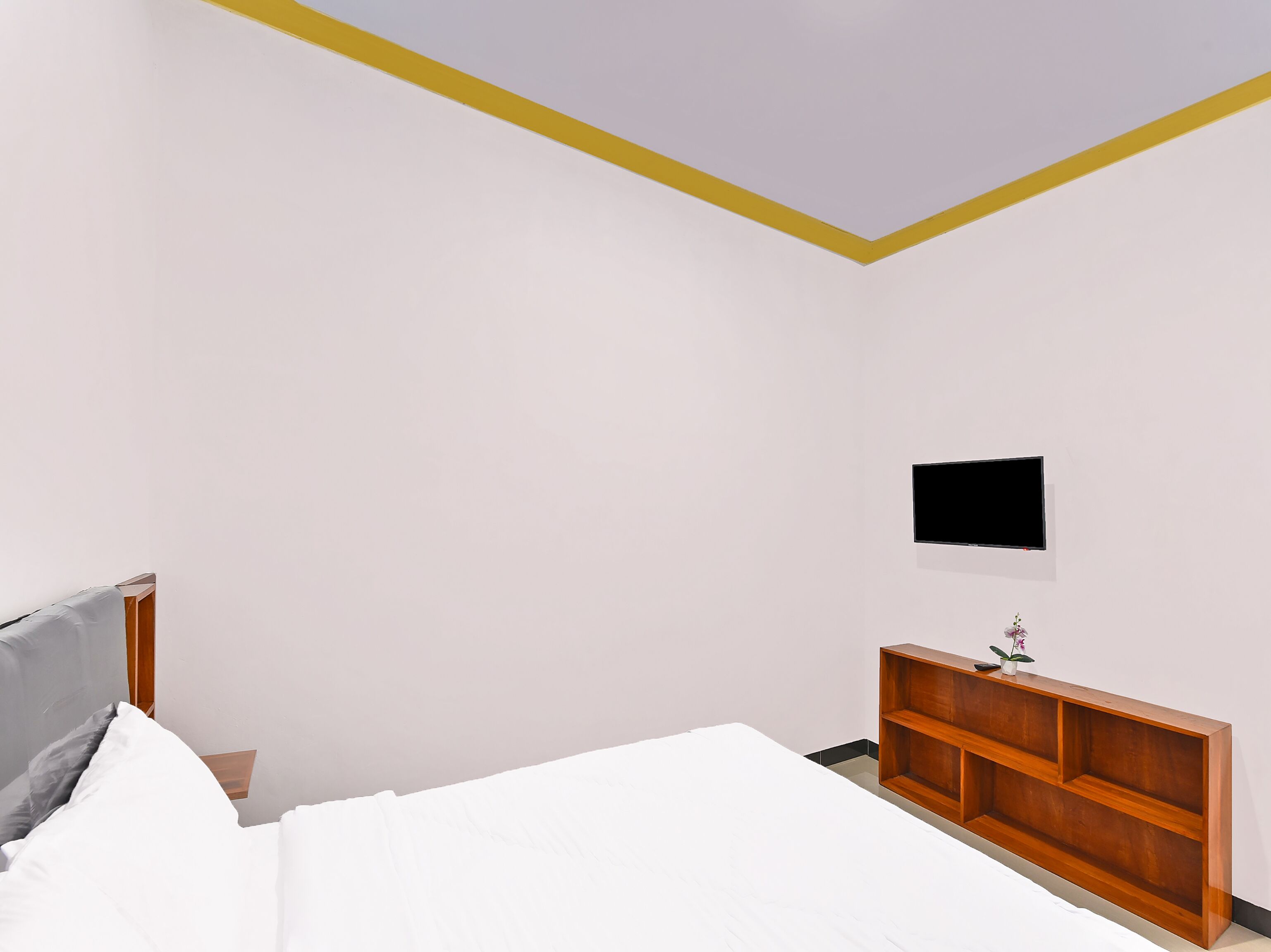 A bedroom with a bed, a wooden nightstand, a TV mounted on the wall, and a small shelf with a plant.