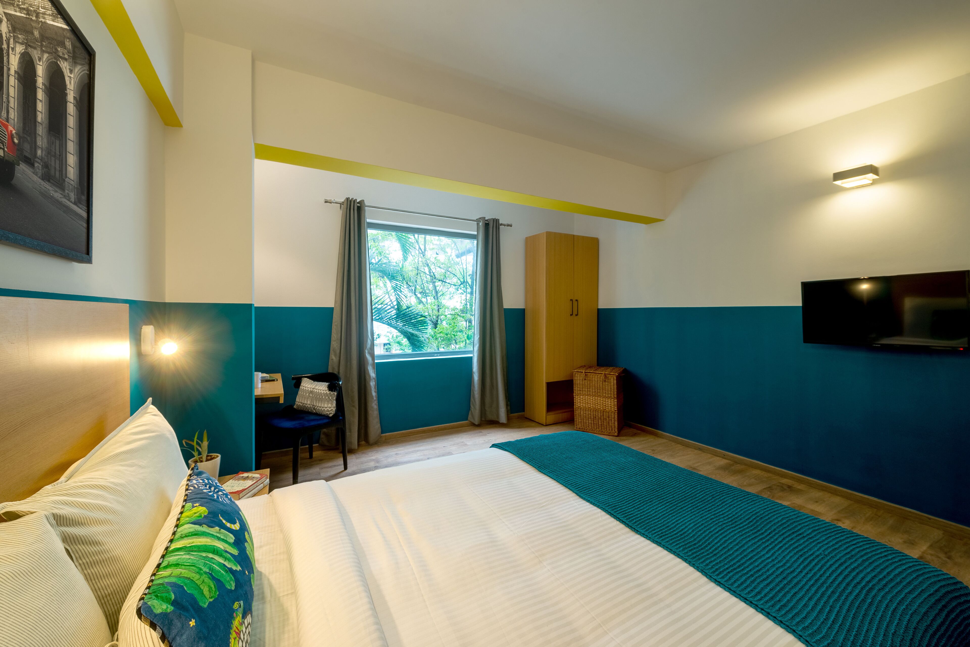 A hotel room with a large bed, a desk with a chair, a TV mounted on the wall, and a window with curtains.