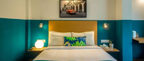 Quarto de hotel com cama, mesa de cabeceira com luminária, quadro na parede e criado-mudo com um livro e uma planta.