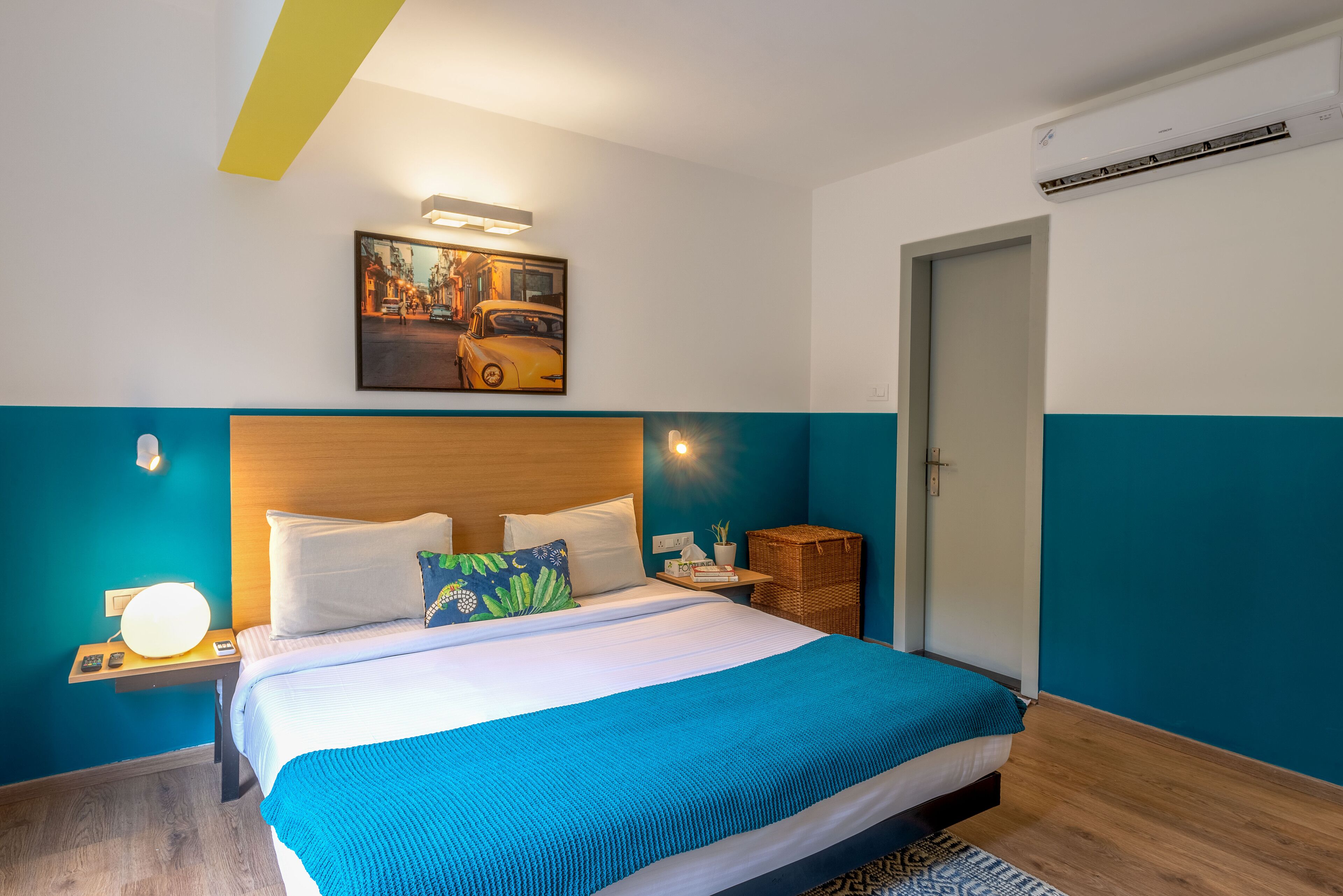 Una habitación de hotel moderna con una cama grande, un cabecero con iluminación integrada, un cuadro colgado en la pared, una mesita de noche con lámpara y una cesta de mimbre.