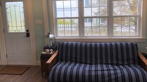 Ein Zimmer mit einer gestreiften Couch, einem kleinen Tisch mit einer Lampe, einer Tür mit einem Buntglasfenster und einem großen Fenster mit Blick auf einen weißen Zaun und Bäume.