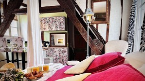 Ein Schlafzimmer mit einem Bett, Kissen, einem Nachttisch mit Lampe sowie einem Frühstückstisch mit Croissants und Orangensaft.