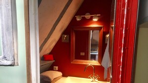 Ein kleines, schmales Badezimmer mit roter Wand, einem weißen Waschbecken und einem Spiegel darüber. Auf dem Waschbecken liegt ein Handtuch und ein rotes Kissen auf einem Regal.