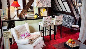 Ein gemütliches Zimmer mit rotem Teppich, einem weißen Sofa mit blumigen Kissen, zwei passenden Sesseln, einem zebra-gemusterten Kissen und einer Holzzahlendecke sowie einem Tisch mit Obst.