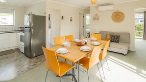 A modern kitchen with a stainless steel refrigerator, a dining table set for four with orange chairs, and a grey sofa in the living area.