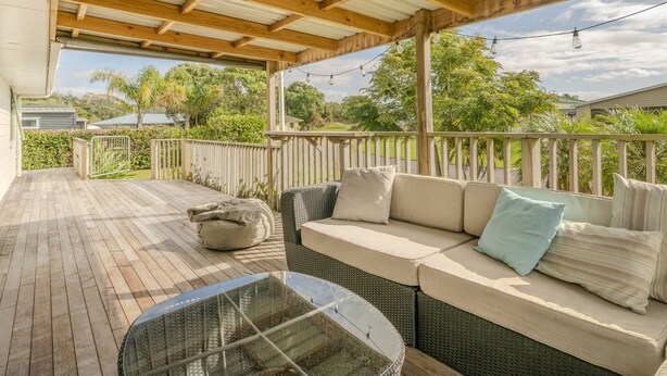 A covered outdoor seating area with a wooden deck, a sofa with cushions, and a glass table.