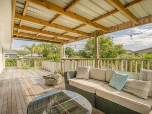 A covered outdoor seating area with a wooden deck, a sofa with cushions, and a glass table. - Sandy Feet Retreat - Cooks Beach Holiday Home (Cooks Beach)
