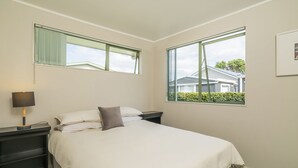 A bedroom with a bed, two bedside tables, a lamp, and two windows with views of houses and greenery.