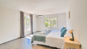 A bedroom with a bed, a bedside table with a lamp, a window with a view of greenery, and a door with curtains. - Modern villa in Las Rotas with air conditioning (Denia)