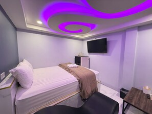 Een moderne slaapkamer met een bed, een bureau, een stoel, een televisie en een prullenbak.