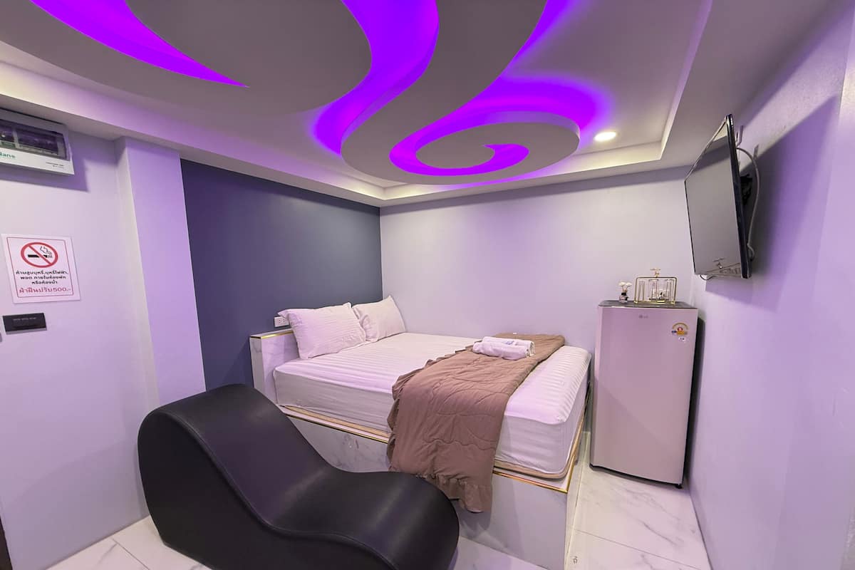 Une chambre avec un lit, une chaise noire, un petit réfrigérateur et un téléviseur fixé au mur.