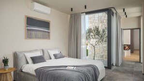 A modern bedroom with a large bed, a view of a stone wall and tree, and a fireplace in the adjacent room.