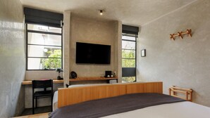 Un dormitorio con una cama grande, un escritorio con una silla, un televisor montado en la pared y una ventana con vistas a zonas verdes.
