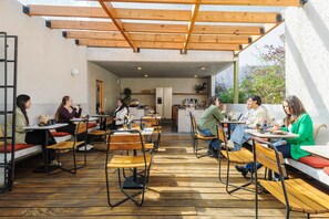 An outdoor cafe with wooden tables and benches, a kitchen area in the background, and a wooden pergola overhead. - Astonishing studio w/terrace @Parque Mexico | Chico (Ciudad de México)