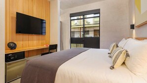 A modern hotel room with a large bed, a flat-screen TV mounted on a wooden panel, a desk with a computer, and a window with a view of buildings. - Amazing studio with Balcony @Parque Mexico | ChiCo (Ciudad de México)