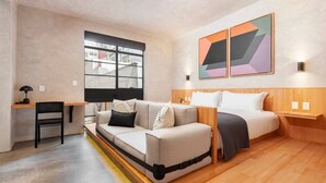 Una habitación moderna con sofá, cama, escritorio y silla. Hay pinturas abstractas en la pared y una ventana con vistas a edificios.