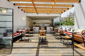 Une terrasse extérieure avec des tables et des bancs en bois, un coin cuisine à l’arrière-plan et une vue sur la verdure.